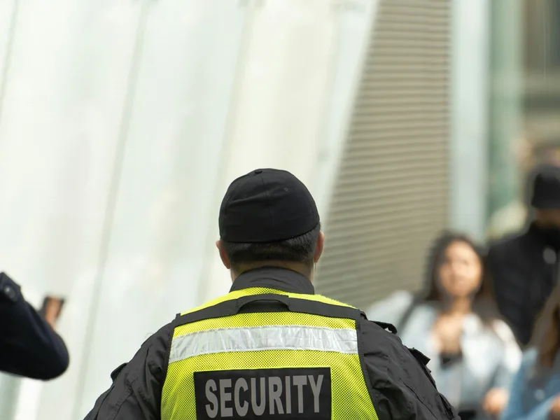 Agent de sécurité privée en uniforme noir et gilet jaune de signalisation, vu de dos, en patrouille devant un bâtiment moderne